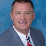 A person in a grey suit with a striped tie and light blue shirt, smiling against a blue background.